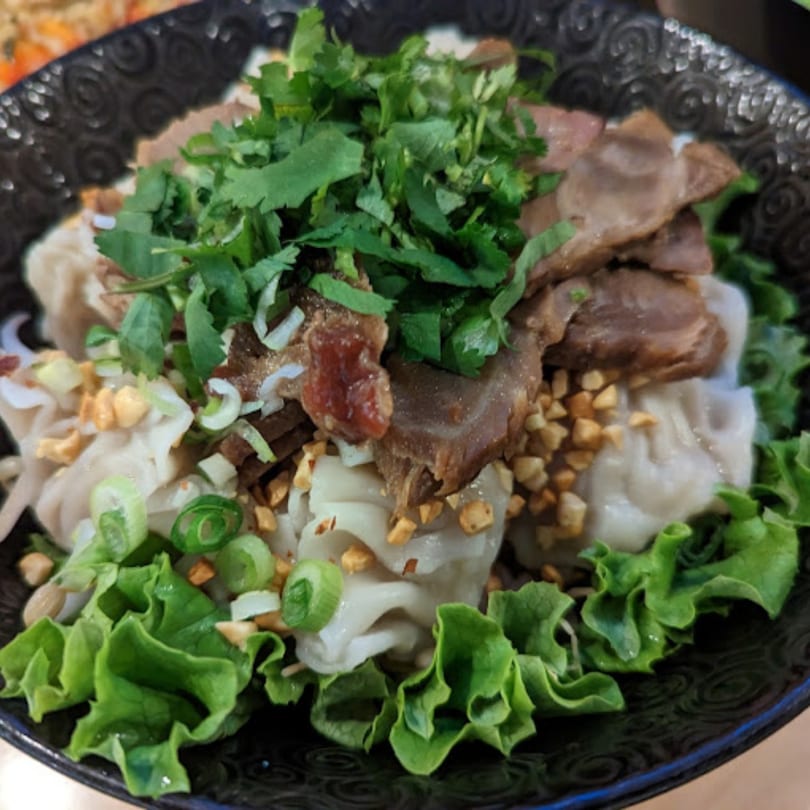 Penny's Oak Park Thai dish with sliced meat, dumplings, lettuce, cilantro, and peanuts in a black bowl.