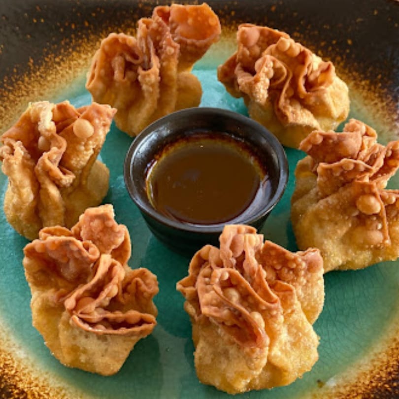 Six crispy fried wontons arranged around a small bowl of dipping sauce on a teal and brown plate.