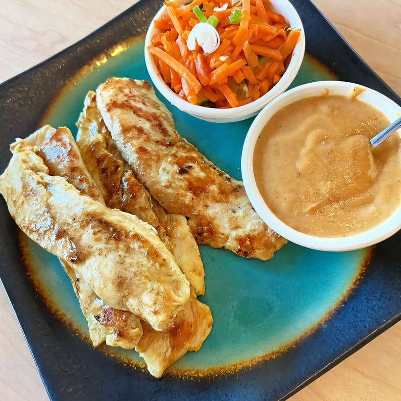 Grilled chicken tenders with peanut sauce and shredded carrots on a blue and black plate.